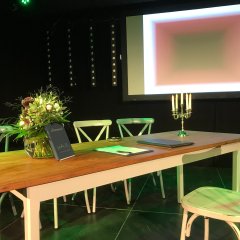 Ein Holztisch mit vier weißen Holzstühlen. Auf dem Tisch befindet sich ein Kerzenständer mit Kerze und ein buntes Blumenbouquet in einer Glasvase, vor der ein schwarzes Stammbuch steht.