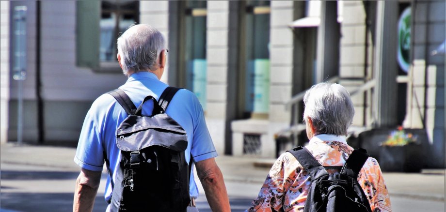 Es ist ein älteres Ehepaar dargestellt die jeweils einen Rucksack aufhaben und spazieren